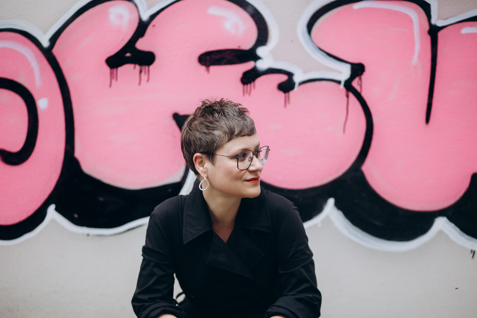 Eine Frau mit kurzem braunem Haar und Brille sitzt vor einer pinken Graffiti-Wand. Sie trägt dunkle Kleidung und silberne Ohrringe und blickt leicht zur Seite.
