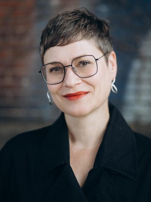 Porträt einer Frau mit kurzem, dunkelbraunem Haar, großer Brille und roten Lippen. Sie trägt silberne Ohrringe und dunkle Kleidung. Der Hintergrund ist weich unscharf und wirkt leicht industriell.